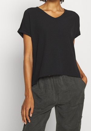 Femme portant un T-shirt noir à manches courtes et col en V, légèrement rentré dans un pantalon taille haute gris foncé, les mains dans les poches.