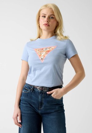 Blondynka w jasnoniebieskim t-shircie z logo GUESS i ciemnoniebieskich dżinsach z brązowym paskiem, stojąca na jednolitym tle.