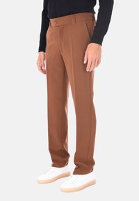 Uomo che indossa pantaloni marroni a gamba dritta, maglietta nera a maniche lunghe e sneakers bianche, in piedi contro uno sfondo neutro.