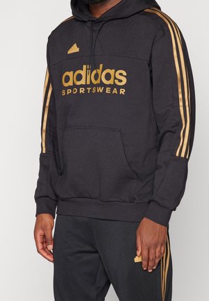 Personne portant un sweat à capuche Adidas noir avec logo et bandes dorées, assorti d'un pantalon noir avec des bandes dorées sur les côtés, debout devant un fond uni.