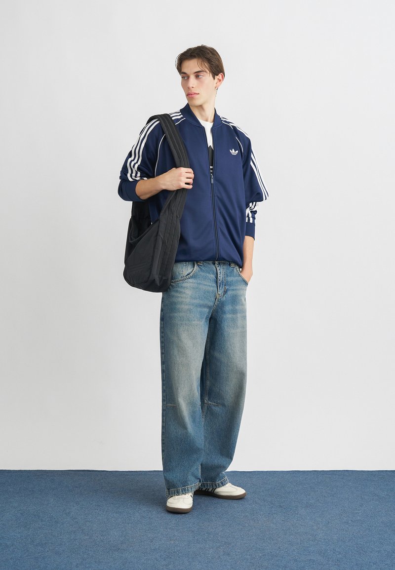 Veste de survêtement zippée bleue marine avec des rayures blanches, jean bleu ample et baskets blanches. Sac à épaule noir. Fond simple et monochromatique.