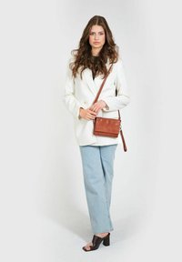 Borsa a tracolla in pelle marrone con superficie texturizzata, hardware argentato e una lunga tracolla regolabile. Indossata con un blazer bianco e jeans azzurri.