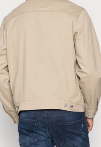 Veste en denim beige avec une coupe ajustée, présentant des détails sur les coutures latérales et des boutons en accent à l'ourlet. Associée à un jean en denim bleu foncé.