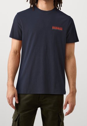 Man wearing navy blue short-sleeve T-shirt with small red and white "Napapijri geographic" logo on left chest and olive cargo pants.