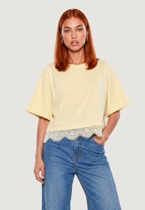 Donna con capelli rossi lisci indossa una maglia gialla pallida ampia con orlo in pizzo e jeans blu a vita alta, con una mano in tasca.