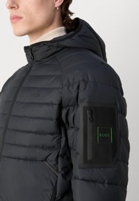 Svart pufferjacka med huva, med horisontellt quiltat mönster, blixtlås-ficka på ärmen och en diskret grön logotyp-accent.