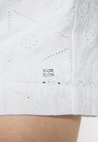 Hvidt broderet stof med blomsterdesign og udskæringsdetaljer. Har et lille metallisk logo-accents på siden.