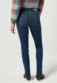 Tajta jeans i mörkblå denim med bakfickor som har zickzack-sömmar. Bärs med en flerfärgad rutig skjorta och svarta skor.