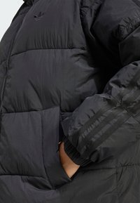 Μαύρο μπουφάν με φερμουάρ, με quilted υφή, πλαϊνές τσέπες και τρεις κάθετες ρίγες στα μανίκια. Περιλαμβάνει ένα μικρό λογότυπο στο στήθος.