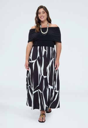 Kvinde smilende, iført en sort off-shoulder top og en lang sort-hvid nederdel med geometrisk mønster samt sorte sandaler.