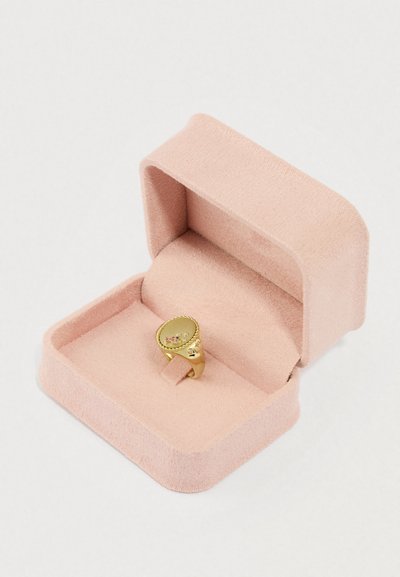 Zlatý prsteň zobrazený v mäkkom ružovom zamatovom puzdre. Má hladký povrch s dekoratívnymi prvkami a malými drahokamami.