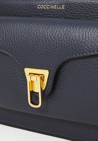 Borsa in pelle blu navy con superficie texturizzata, hardware dorato e forma strutturata, caratterizzata da un evidente dettaglio della chiusura. Nome del brand impresso.