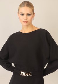 Czarny prążkowany top z długim rękawem i szerokim dekoltem oraz oversize'owymi rękawami, przewiązany w talii złotym łańcuszkowym paskiem.