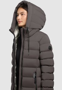 Giubbotto puffer grigio scuro con un grande cappuccio regolabile. Caratterizzato da una fodera interna lucida e tasche con zip. Stringhe regolabili sul cappuccio.