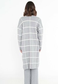 Long gilet gris avec un motif à carreaux blancs, présentant des épaules tombantes et une coupe droite. Porté par-dessus un pantalon à côtes gris clair.