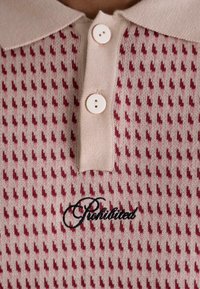 Beige knit polo shirt featuring a collared neckline, two white buttons, a textured pattern of small red accent shapes, and embroidered "Prohibited."