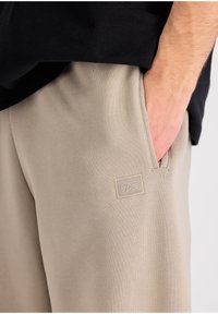 Beige trenirke iz mehkega blaga z teksturirano površino. Imajo stranski žep in majhen pravokoten logotip.