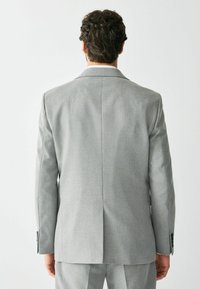 Hombre con cabello rizado oscuro, vestido con un traje gris claro, de pie y de espaldas contra un fondo blanco liso.