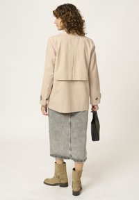 Chaqueta beige corta con un pliegue en la parte posterior, combinada con una falda de mezclilla gris y botas de ante marrón. Un bolso negro completa el conjunto.