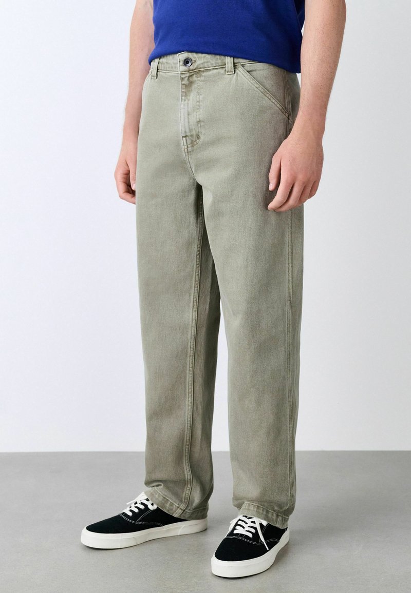 Light green, straight-leg jeans feature a button closure, front pockets, and visible stitching. Paired with black sneakers and a blue shirt.