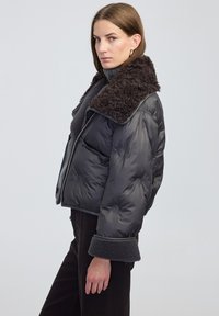 Kobieta w czarnej pikowanej kurtce z dużym fakturowanym kołnierzem, podwiniętymi mankietami i czarnymi spodniami, stojąca na tle jasnego, jednolitego tła.