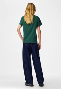 Persoon met lang blond haar, gekleed in een groen shirt met korte mouwen, donkerblauwe wijde jeans en zwarte sneakers, staand met de rug naar de kijker toe.