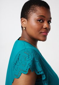 Blouse teal avec des manches courtes en dentelle détaillées et un col rond. Texture lisse, avec broderies et motifs découpés.