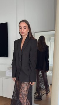 Črni blazer z enim gumbom in dvema žepoma, kombiniran s prosojno črno čipkasto krilo, ki prikazuje cvetlične vzorce in tekoč dizajn.