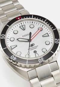 Montre en acier inoxydable avec un cadran blanc, une lunette noire et des accents rouges. Dispose d'index horaires lumineux et d'une étanchéité allant jusqu'à 200 m.