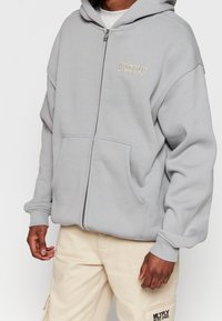 Hellgrauer Kapuzenpullover mit Reißverschluss und Fronttasche, mit gesticktem Text auf der linken Seite und gerippten Bündchen für eine eng anliegende Passform.