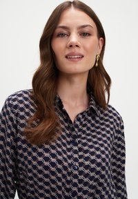 Granatowa koszula z geometrycznym wzorem w białym i jasnoróżowym kolorze, zapinana na guziki, z kołnierzykiem, z niebieskimi guzikami i miękką teksturą materiału.