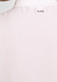 Chemise rose clair à texture lisse, avec un col classique et une étiquette logo argentée à l'arrière près de l'encolure.