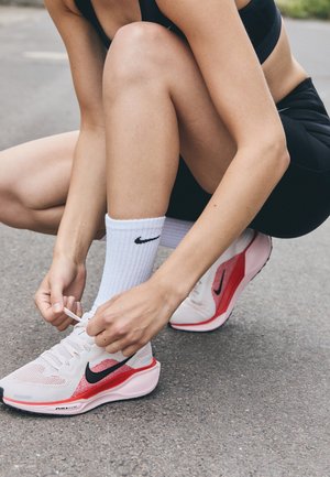 Nike running shoes with a pink and white mesh upper, black accents, and a textured sole. A person ties the laces while squatting.