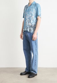 Koszula z krótkim rękawem w jasnoniebieskim kolorze z koralowym wzorem, zestawiona z jasnoniebieskimi dżinsami i czarnymi mokasynami, stojąca na neutralnym tle.