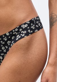 Κοντινό πλάνο στο ισχίο και το πάνω μέρος του μηρού ενός ατόμου που φοράει μαύρο εσώρουχο με λουλουδάτο σχέδιο, με ένα τατουάζ στο χέρι.