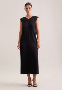 Robe noire sans manches avec un décolleté en V, une silhouette droite et des coutures minimales. Associée à des talons à bride à la cheville. Texture lisse.