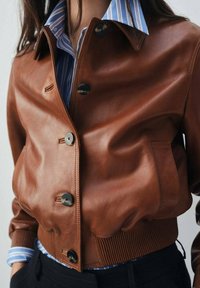 Veste en cuir brun courte avec des boutons noirs, un ourlet côtelé et une chemise à rayures bleues en dessous. Texture lisse, design ajusté.