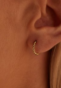 Boucle d'oreille en or, design tordu, petite taille, présentant une texture lisse et une fermeture sans couture, portée dans le lobe de l'oreille.