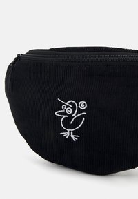 Svart corduroy veske med rund form, med et hvitt brodert fuglemotiv og glidelås på toppen. Teksturert overflatedetalj synlig.