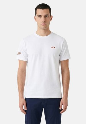 Uomo che indossa una t-shirt bianca a maniche corte con piccoli simboli marroni sul petto e sulla manica sinistra, abbinata a pantaloni blu navy, in piedi immobile.