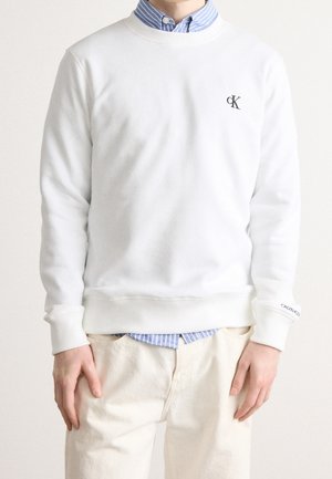 Mann trägt ein weißes Calvin Klein Sweatshirt über einem blau gestreiften Hemd und beigen Hosen, steht mit locker an den Seiten hängenden Händen.