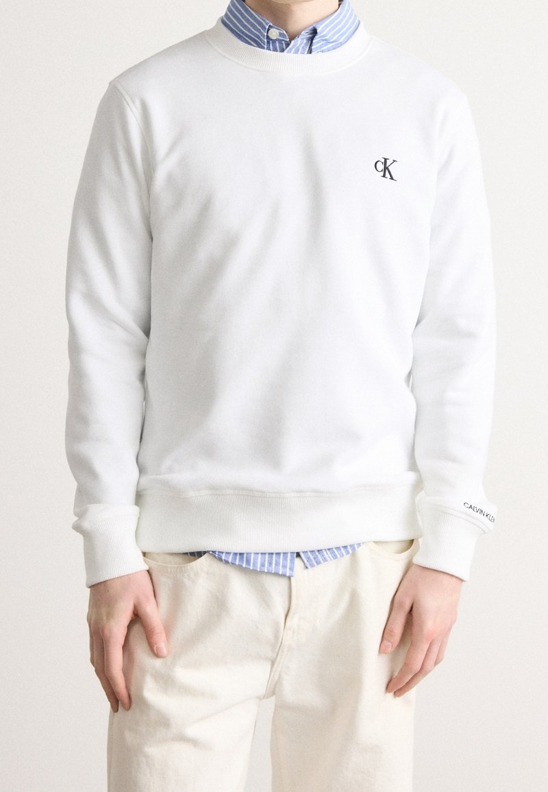 Homme portant un sweat-shirt blanc Calvin Klein par-dessus une chemise à rayures bleues boutonnée et un pantalon beige, debout les mains détendues le long du corps.