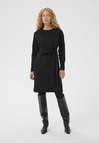 Czarna sukienka z długim rękawem o dopasowanym kroju, z cechą nawiązującą do owijania oraz wiązania w talii, wykonana z miękkiego, rozciągliwego materiału.