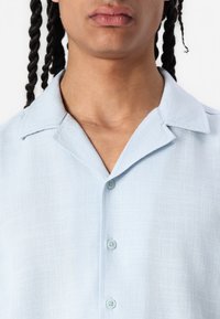 Chemise courte à manches courtes, bleu clair, avec un col, comportant cinq boutons, en tissu texturé, et un subtil motif à carreaux.