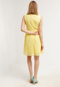 Vestido amarillo sin mangas con patrón perforado, cintura entallada y falda acampanada. Combinado con sandalias de tacón azul claro.