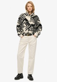 Jonge vrouw in een zwart-wit gestreepte fleece pullover, witte high-waist broek met een zwarte riem en zwarte veterschoenen.