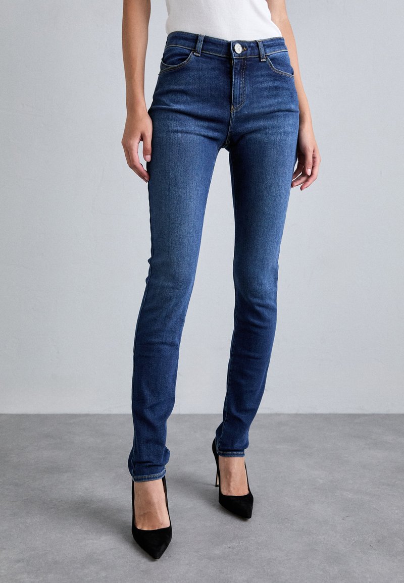 Smala jeans i mörkblå denim med en slät yta, femfickorsdesign, knappstängning och en figurnära silhuett. Kombineras med svarta klackar.