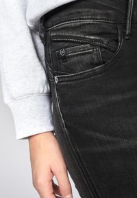 Jean en denim noir avec une coupe slim. Les caractéristiques incluent un léger délavage, une poche avant et une petite étiquette logo. Associé à un sweat-shirt gris avec des poignets.