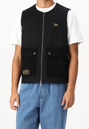 Mann trägt eine schwarze Zip-up-Weste mit zwei vorderen Klappentaschen über einem weißen T-Shirt und lockeren blauen Jeans, steht mit Armen an den Seiten.