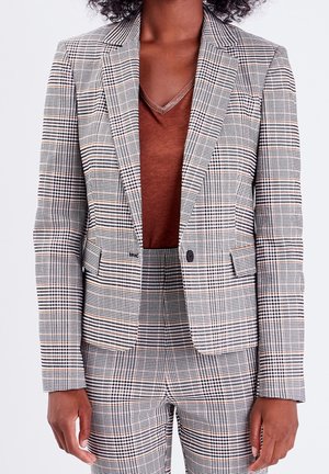 Femme portant un haut marron à col en V sous un blazer à carreaux gris et noir et pantalon taille haute assorti.